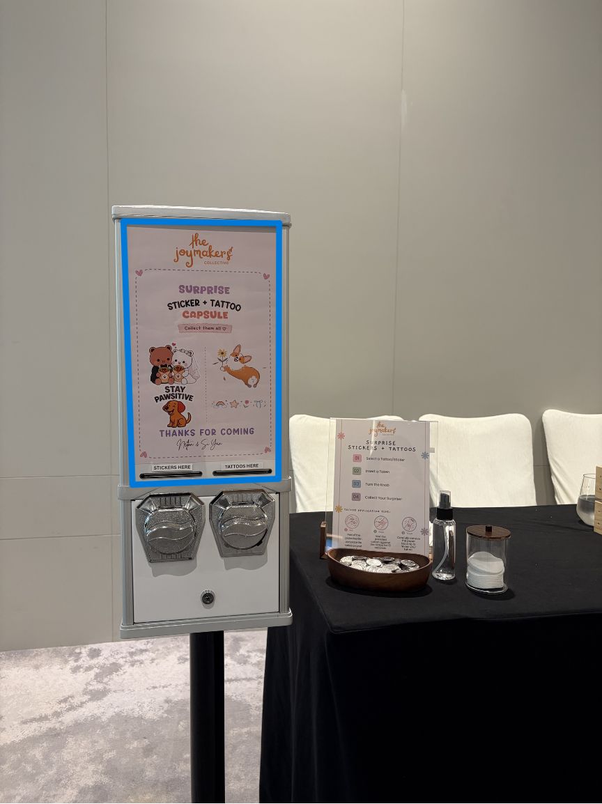 The Keepsake Bar's sticker and tattoo capsule machine beside a table with a vanity spray, at a hotel event
