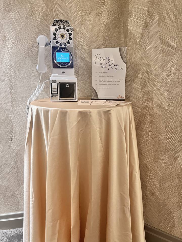 A white retro payphone guestbook and wedding sign on a round table with a floor-length champagne cloth